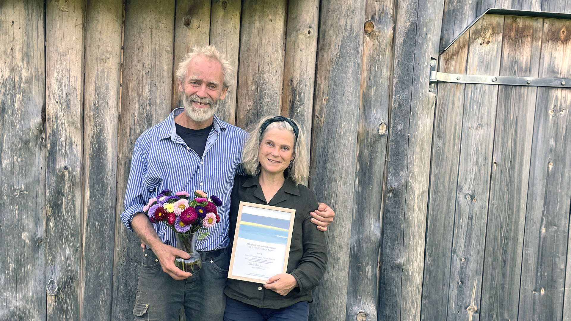 Kvinna och man framför står framför gammal ladugårdsvägg, med diplom och blommor